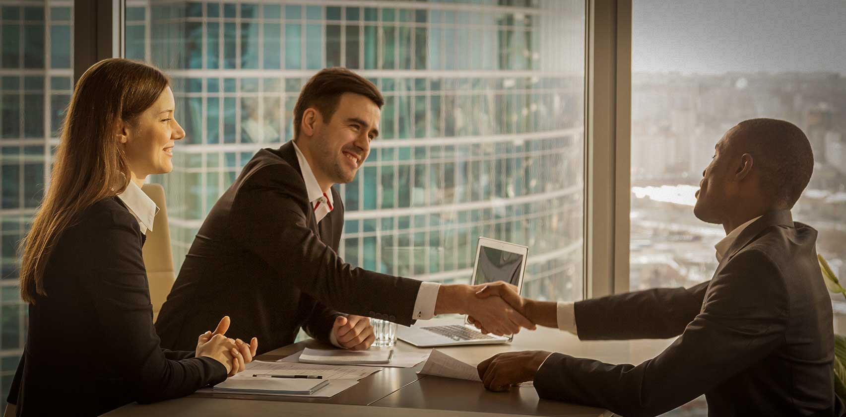 Three people sat around an office table shaking hands with smiles on their face.