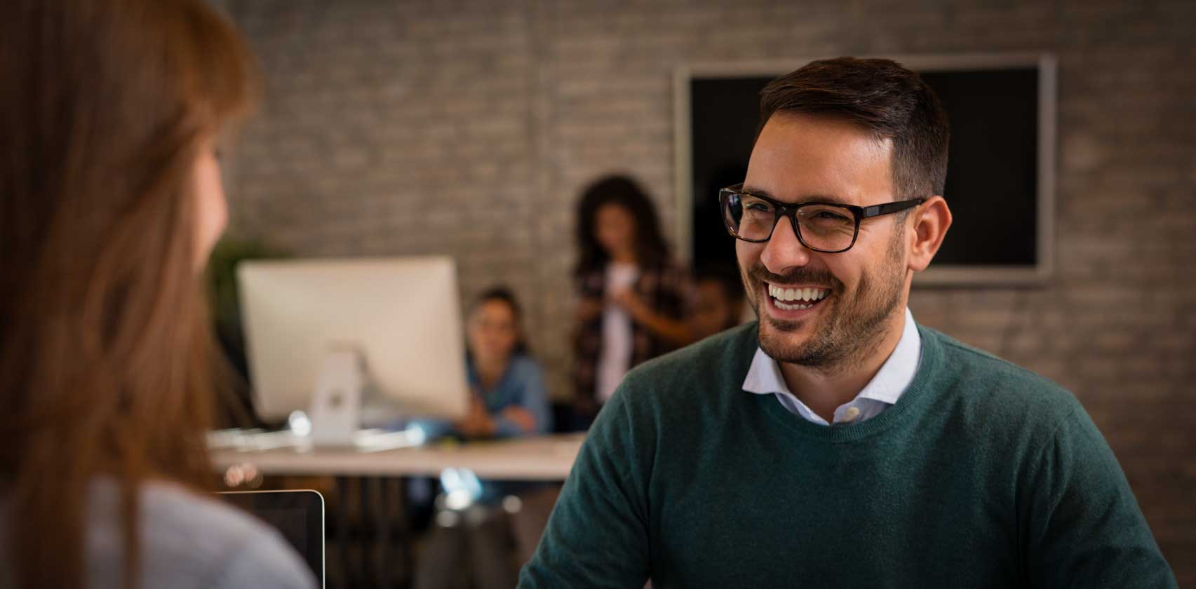 Main focus of a smiling man with glasses speaking to a women who is in soft focus nearer the camera.