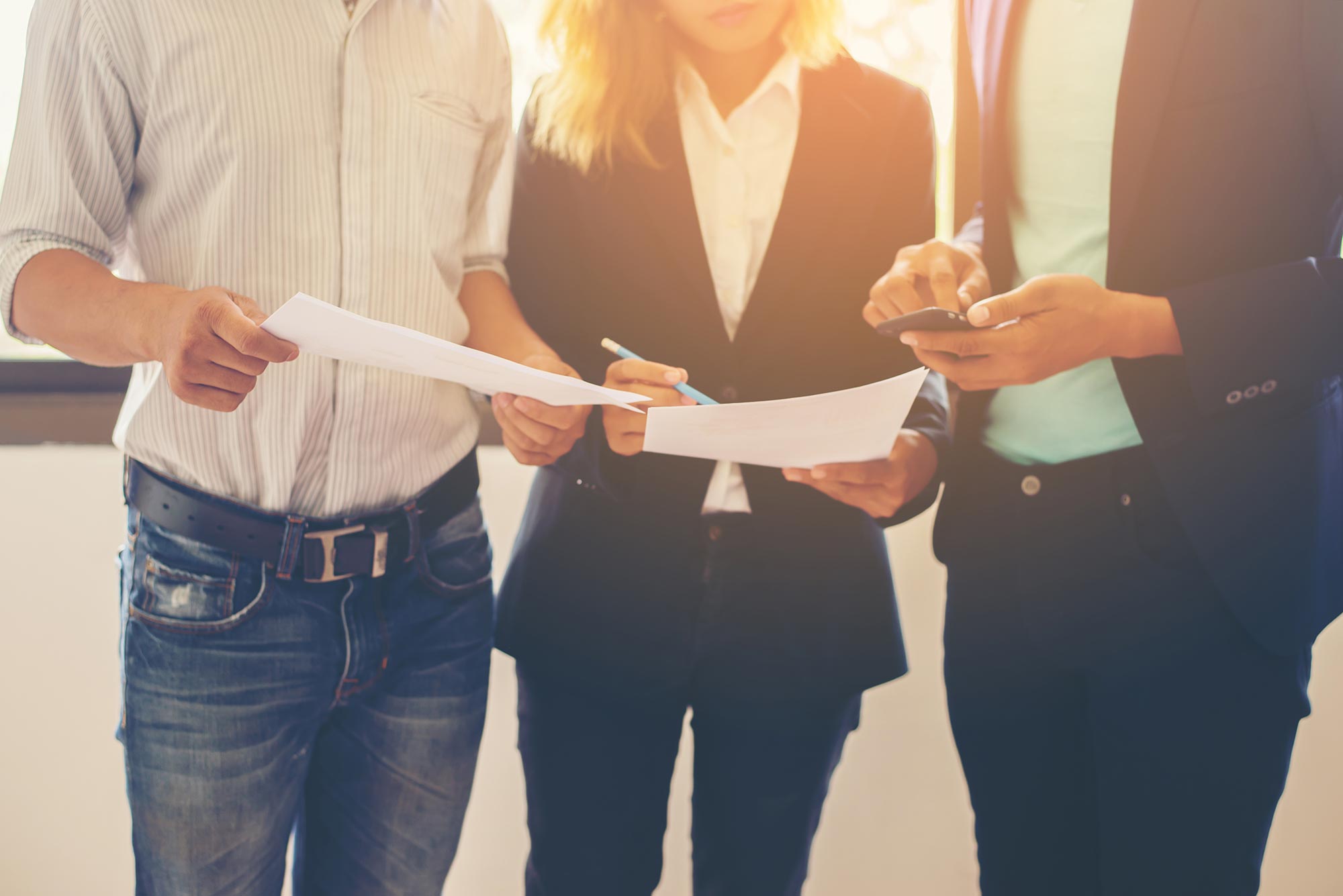 Close up of three people stood up two of them holding paperwork and the other writing something down on their phone.